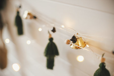 Stylish christmas garland close up with bells and trees on white wooden wall with christmas lights. Festive decorated atmospheric scandinavian room. Modern boho handmade decor. Space for textの写真素材