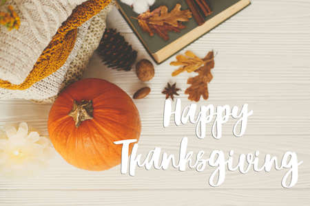 Happy thanksgiving text on pumpkin, cozy sweaters, autumn leaves, candle and vintage book on white wood flat lay. Seasonal greeting card, handwritten sign. Give thanksの写真素材