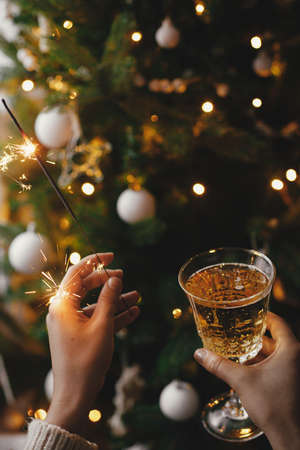 Hands holding burning sparkler and champagne glass on background of christmas tree lights. Happy New Year! Firework bengal light and drink in woman hands. Atmospheric moment. New Year eve partyの写真素材