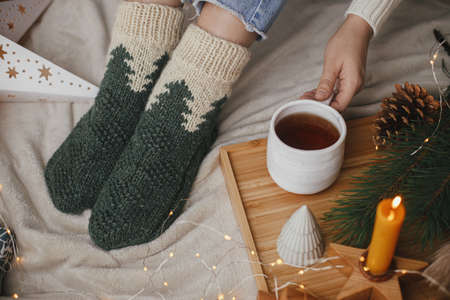 Cozy winter moments at home. Woman feet in woolen socks on soft bed with warm cup of tea, christmas stars, golden lights, trees, candle. Girl relaxing in scandinavian evening roomの写真素材
