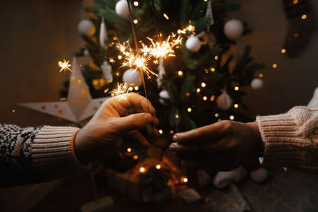 Couple celebrating with firework bengal lights on background of christmas tree and glowing star. Happy New Year! Hands holding burning sparklers in festive scandinavian room. Atmospheric momentの写真素材