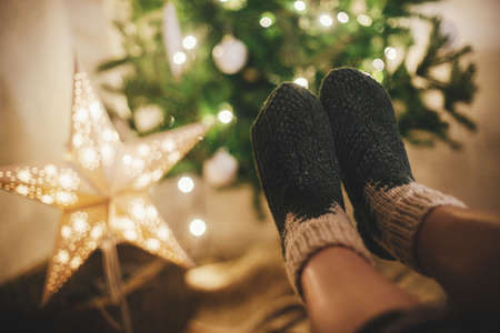 Woman feet in cozy woolen socks on background of christmas tree and star in lights in festive evening room. Cozy winter moments at home. Stylish warm socks on female legsの写真素材