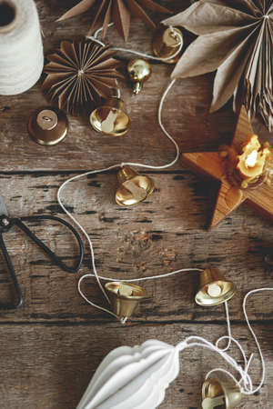 Stylish Christmas bells, paper angel and stars, ornaments, candle, thread and scissors on rustic wooden background. Making festive scandinavian garland, moody atmospheric imageの写真素材