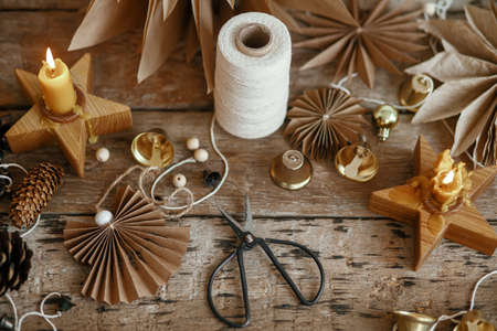 Stylish Christmas bells, paper angel and stars, pine cones, candle, thread and scissors on rustic wooden background. Making festive scandinavian garland, moody image. Handmade holiday decorの写真素材