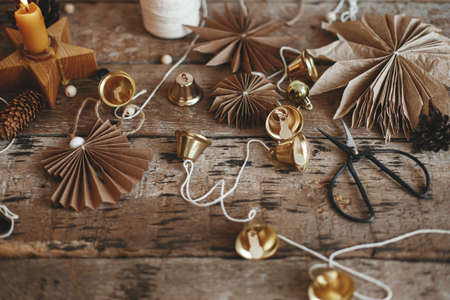 Stylish Christmas bells, paper angel and stars, candle, thread and scissors on rustic wooden background. Making festive scandinavian garland, atmospheric holiday decor. Merry Christmasの写真素材