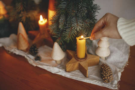 Hand lighting up christmas candle on background of pine trees decorations, cones and rustic cloth on wooden table in evening scandinavian festive room. Holiday advent. Atmospheric winter timeの写真素材