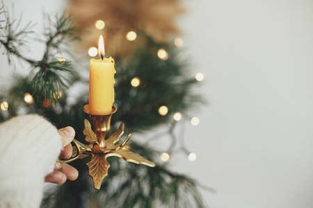 Atmospheric hygge winter home. Hand in cozy sweater holding vintage candlestick with burning candle on background of warm lights, fir branches, sweden star in festive scandinavian roomの写真素材