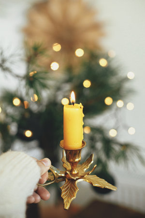 Hand in cozy sweater holding vintage candlestick with burning candle on background of warm lights, fir branches, sweden star in festive scandinavian room. Atmospheric hygge home. Christmas adventの写真素材