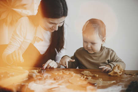 Adorable little daughter and mother making together gingerbread cookies on messy wooden table in...