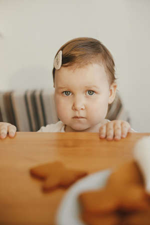 Cute little girl looking at freshly baked gingerbread cookie on wooden table close up. Authentic lovely moment, holiday preparations. Adorable toddler girl making christmas cookies with motherの写真素材