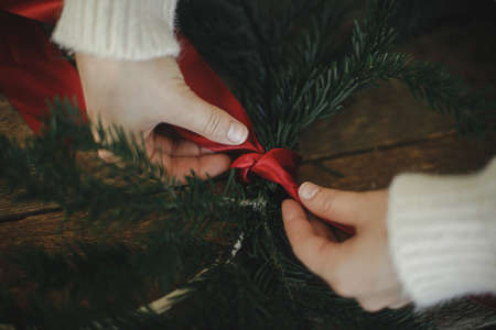 Hands making modern christmas wreath with fir branches and red ribbon on rustic table. Atmospheric moody image. Winter holidays preparation. Making minimalist xmas wreathの写真素材
