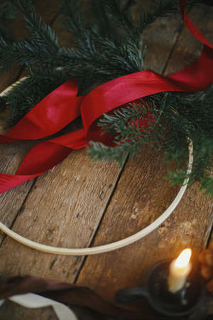 Making stylish modern christmas wreath. Fir branches, round wooden hoop, red ribbon, candle and pine cones on rustic table. Atmospheric moody image. Winter holidays preparationの写真素材