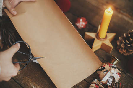Wrapping christmas gift. Hands cutting stylish wrapping paper with scissors on rustic wooden table background with candle and red ornaments. Atmospheric image. Winter Holidays preparationの写真素材