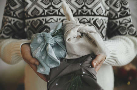 Zero waste and eco friendly presents, Christmas Furoshiki gifts. Woman hands in cozy sweater holding wrapped xmas gift in fabric in scandinavian room. Atmospheric moody image, nordic style.の写真素材