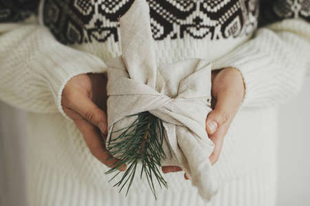 Christmas Furoshiki gift. Christmas gift wrapped in fabric in hands of woman in cozy sweater in white scandinavian room. Atmospheric moody image. Zero waste and eco friendly presentsの写真素材