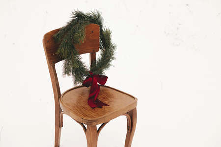 Merry Christmas! Christmas wreath on rustic chair in snowy winter field. Winter holidays in countryside. Space for text. Stylish xmas wreath with pine branches and red bow hanging on wooden chairの写真素材