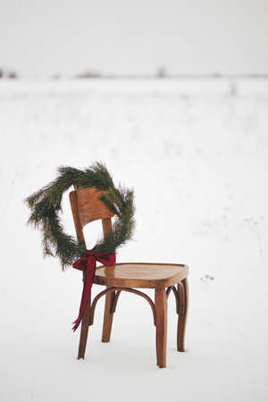 Merry Christmas! Christmas wreath on rustic chair in snowy winter field. Winter holidays in countryside. Stylish xmas wreath with pine branches and red bow hanging on wooden chair. Atmospheric timeの写真素材