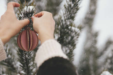 Hands in cozy sweater holding stylish ornament on background of pine tree branches in snow....