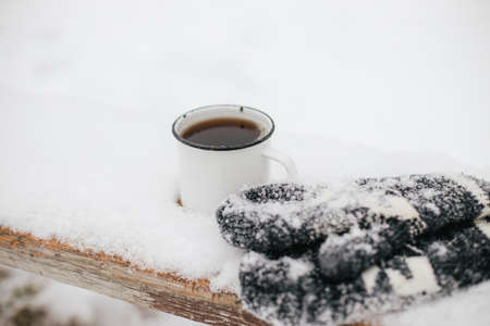 Warm cup of tea and cozy knitted mittens in snow on wooden bench in winter park. Hiking and traveling in cold season. Wanderlust. Space for text. Winter timeの写真素材