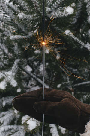 Happy New Year!  Woman hand in stylish mitten with glowing firework on background of pine tree branches in snow in evening. Hand holding burning sparkler. Atmospheric magic moment. Happy Holidays!の写真素材