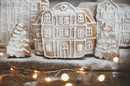 Merry Christmas and Happy Holidays! Christmas gingerbread houses and trees in snow on rustic wood with golden lights. Atmospheric moody image. Christmas cookies village scene. Season greetingの写真素材