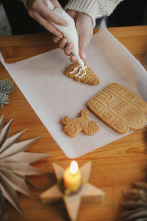 Hands decorating christmas gingerbread cookies with icing on wooden table with candle and ornaments. Close up. Making gingerbread house with frosting. Holiday preparation and traditionの写真素材