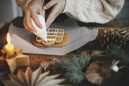 Hands decorating christmas gingerbread cookies with icing on wooden table with candle and ornaments. Close up. Making gingerbread house with frosting. Holiday preparation and traditionの写真素材