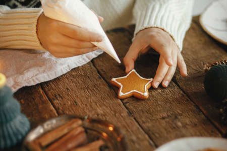 Hands decorating christmas gingerbread cookie star with icing on rustic table with candle, spices, decorations. Atmospheric moody image. Woman making traditional christmas gingerbread cookiesの写真素材