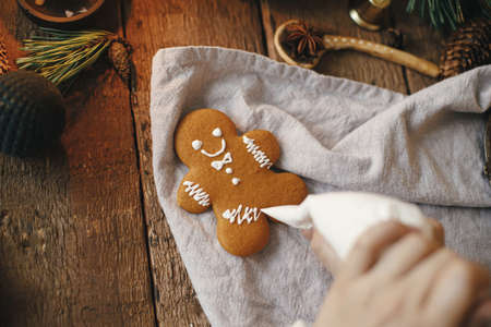 Hands decorating christmas gingerbread man cookie with frosting on rustic table with napkin, spices, decorations. Atmospheric moody time. Making traditional christmas gingerbread cookiesの写真素材
