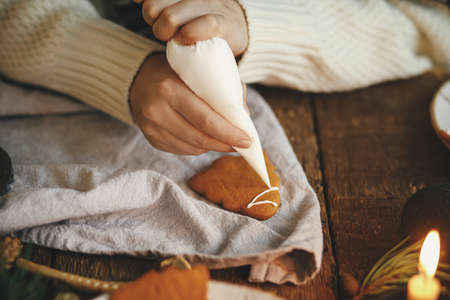 Hands decorating christmas gingerbread cookie tree with frosting on rustic table with napkin, candle, decorations. Atmospheric moody time. Making traditional christmas gingerbread cookiesの写真素材