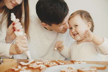 Happy stylish family decorating christmas gingerbread cookies with icing on wooden table. Cute little daughter, mom and dad time together. Xmas holiday preparations. Authentic lovely momentsの写真素材