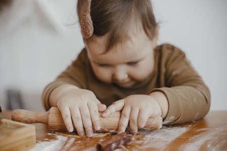 Adorable baby girl kneading dough with wooden rolling pin for gingerbread cookies on messy table, hands close up. Cute toddler daughter helps mother making christmas cookies. Authentic momentsの写真素材
