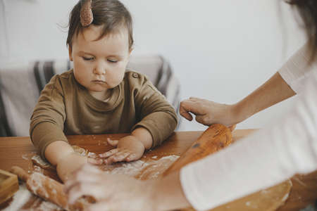 Adorable baby daughter kneading dough together with mother for gingerbread cookies on wooden table in modern scandinavian room. Cute toddler girl helping making christmas cookies. Authentic momentsの写真素材