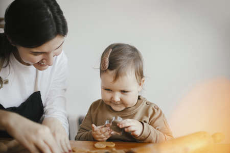 Adorable little daughter with mother making together christmas gingerbread cookies on messy wooden table. Cute toddler girl helps cutting dough with festive cutters for cookies. Family timeの写真素材