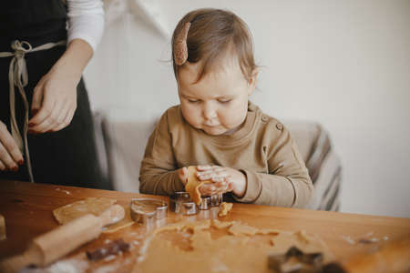 Adorable little daughter with mother making together christmas gingerbread cookies on messy wooden table. Cute toddler girl helps cutting dough with festive cutters for cookies. Family timeの写真素材