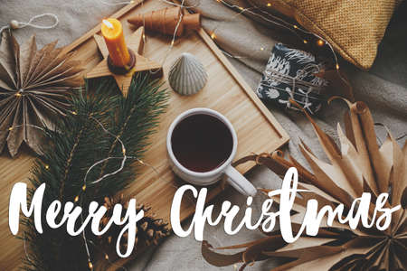 Merry Christmas greeting card. Merry Christmas text on warm cup of tea, christmas lights, stars, pine trees, candle flat lay. Winter hygge. Happy Holidays! Handwritten signの写真素材