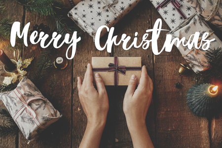 Merry Christmas text on hands holding stylish christmas gift wrapped in craft paper on rustic table flat lay. Season's greetings card. Handwritten sign. Happy Holidays!の写真素材