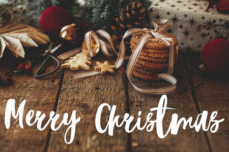 Merry Christmas greeting card. Merry Christmas text on cookies, stylish gift, festive decorations on rustic table. Atmospheric xmas composition. Happy Holidays! Handwritten signの写真素材