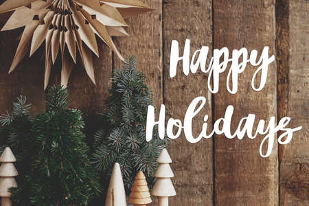 Happy Holidays text on stylish christmas paper star and miniature pine trees on rustic background. Season's greetings card. Handwritten sign. Merry Christmas!