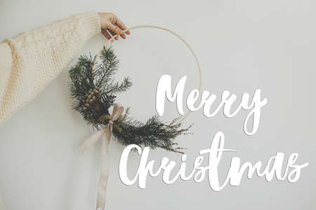 Merry Christmas text on hand holding modern boho christmas wreath on white background. Season's greetings card. Handwritten sign. Happy Holidays!の写真素材