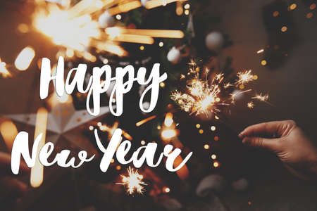 Happy New Year text on hands holding burning sparklers on background of christmas tree in lights. Season's greetings card. Handwritten sign. Happy holidaysの写真素材