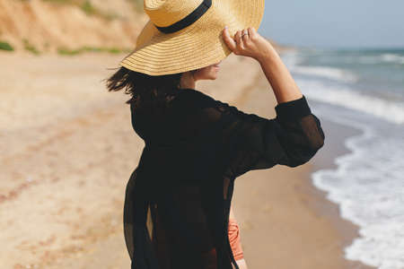 Stylish carefree woman in hat walking on sunny beach at sea and enjoying summer vacation. Young...