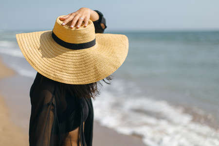 Stylish carefree woman holding hat and relaxing on sunny beach at sea, close up. Summer vacation. Young fashionable female in straw hat and light shirt enjoying holiday on tropical islandの写真素材