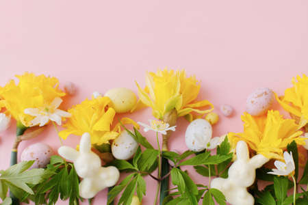 Happy Easter! Colorful Easter chocolate eggs, bunnies and daffodils flowers border on pink background. Stylish Easter flat lay. Greeting card or banner templateの写真素材