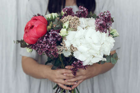 Woman in rustic dress holding beautiful peony, lilac, roses, hydrangea flowers and eucalyptus...
