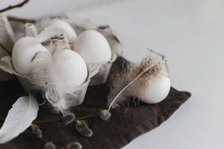 Easter simple aesthetics. Natural eggs in tray, feathers, willow branches on rustic linen napkin on white wooden table. Stylish rural Easter still life in pastel white and brown colorsの写真素材