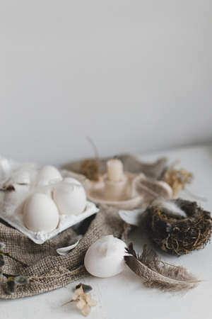 Easter rustic still life. Easter natural eggs in tray, feathers, willow branches, nest on burlap on white wooden table. Simple rural Easter aesthetics in pastel beige colors. Happy Easterの写真素材