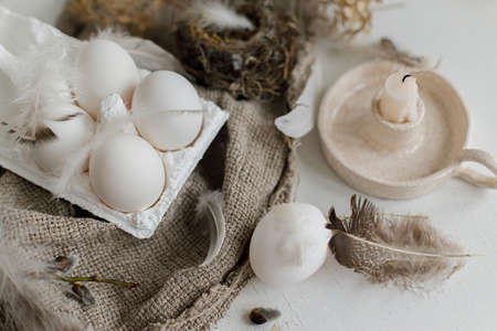 Easter rustic still life. Easter natural eggs in tray, feathers, willow branches, nest on burlap on white wooden table. Simple rural Easter aesthetics in pastel beige colors. Happy Easterの写真素材