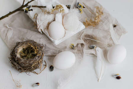 Easter natural eggs, feathers, willow branches, nest on rustic white table. Stylish rural Easter still life in pastel white and beige colors. Modern simple aestheticsの写真素材