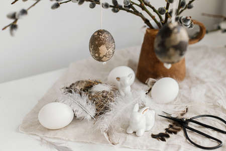 Easter rustic still life. Natural eggs, bunny figurines, feathers, nest, willow branches in vase on aged wooden table. Simple stylish easter decoration on table. Happy Easterの写真素材